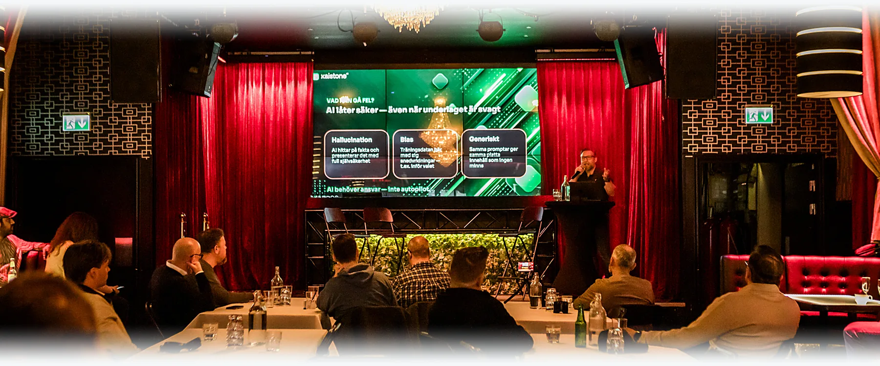 A speaker on stage in front of a large screen and red curtains, the audience at the tables.